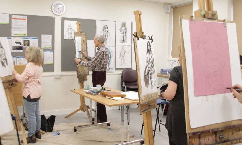 4 people in a classroom with easels drawing a live model