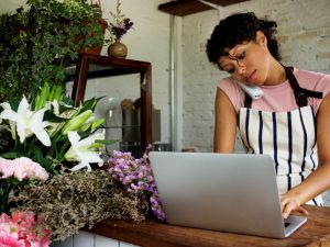 Lady wearing pink tshirt and stripy apron on telephone and typing at a laptop in a florist