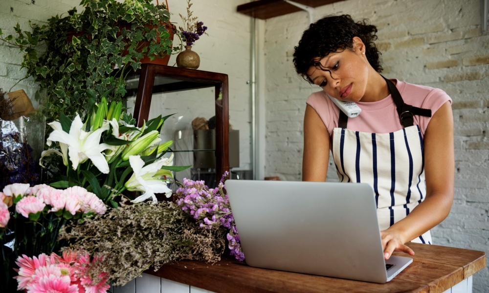 Lady wearing pink tshirt and stripy apron on telephone and typing at a laptop in a florist